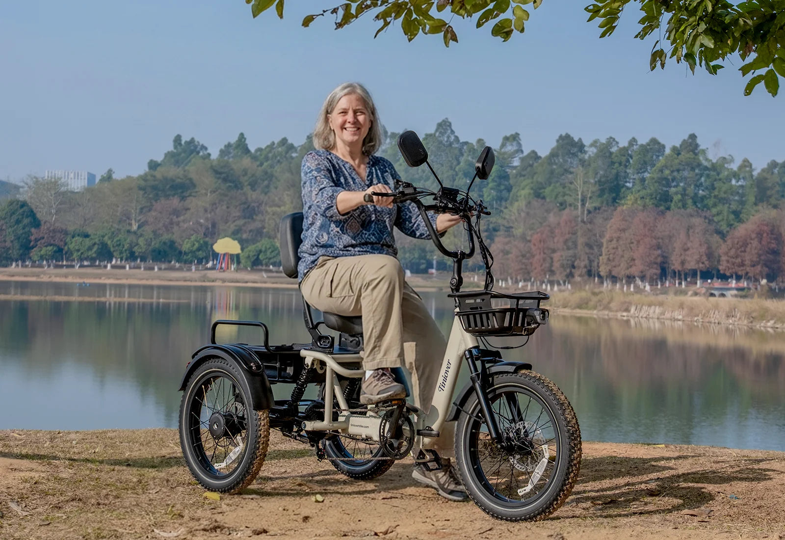White Tiniover River electric trike with a rider, front basket, rear suspension, and lakeside background