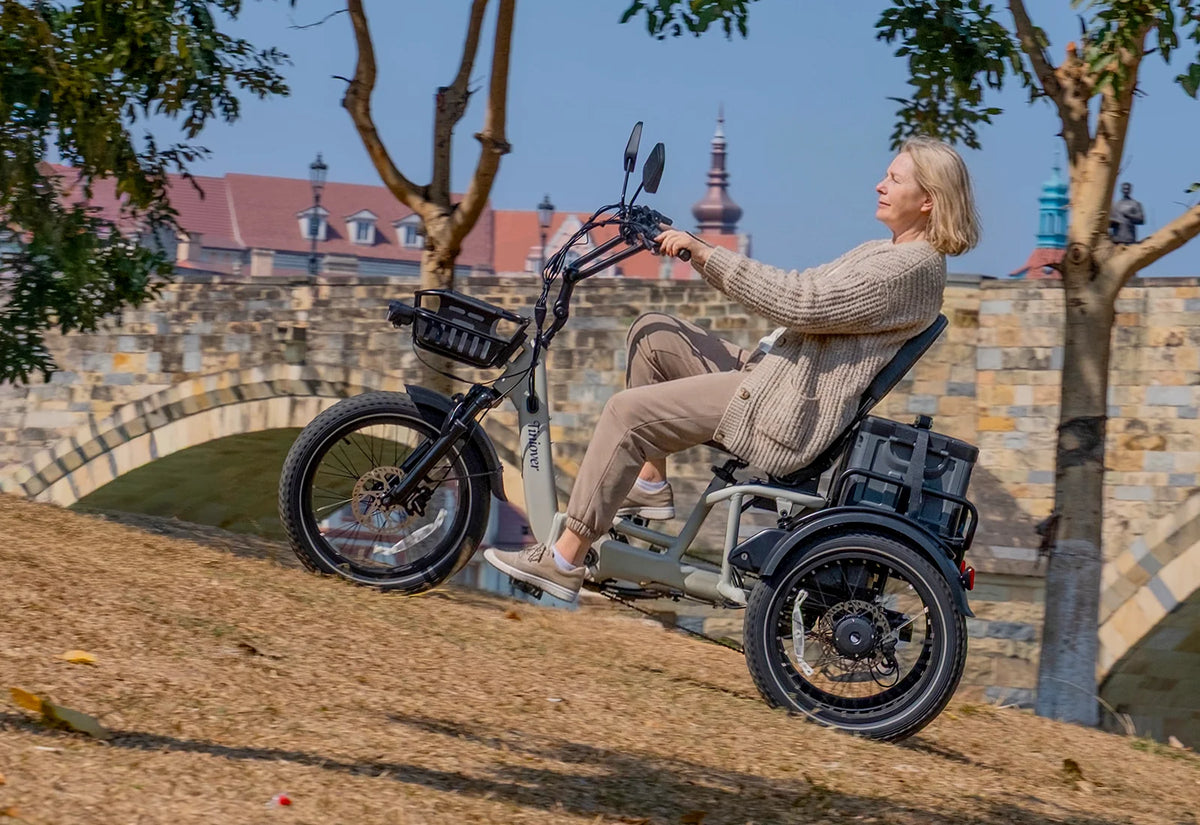 White Tiniover River electric trike with a rider, front basket, rear storage box, and fat tires on a sloped path