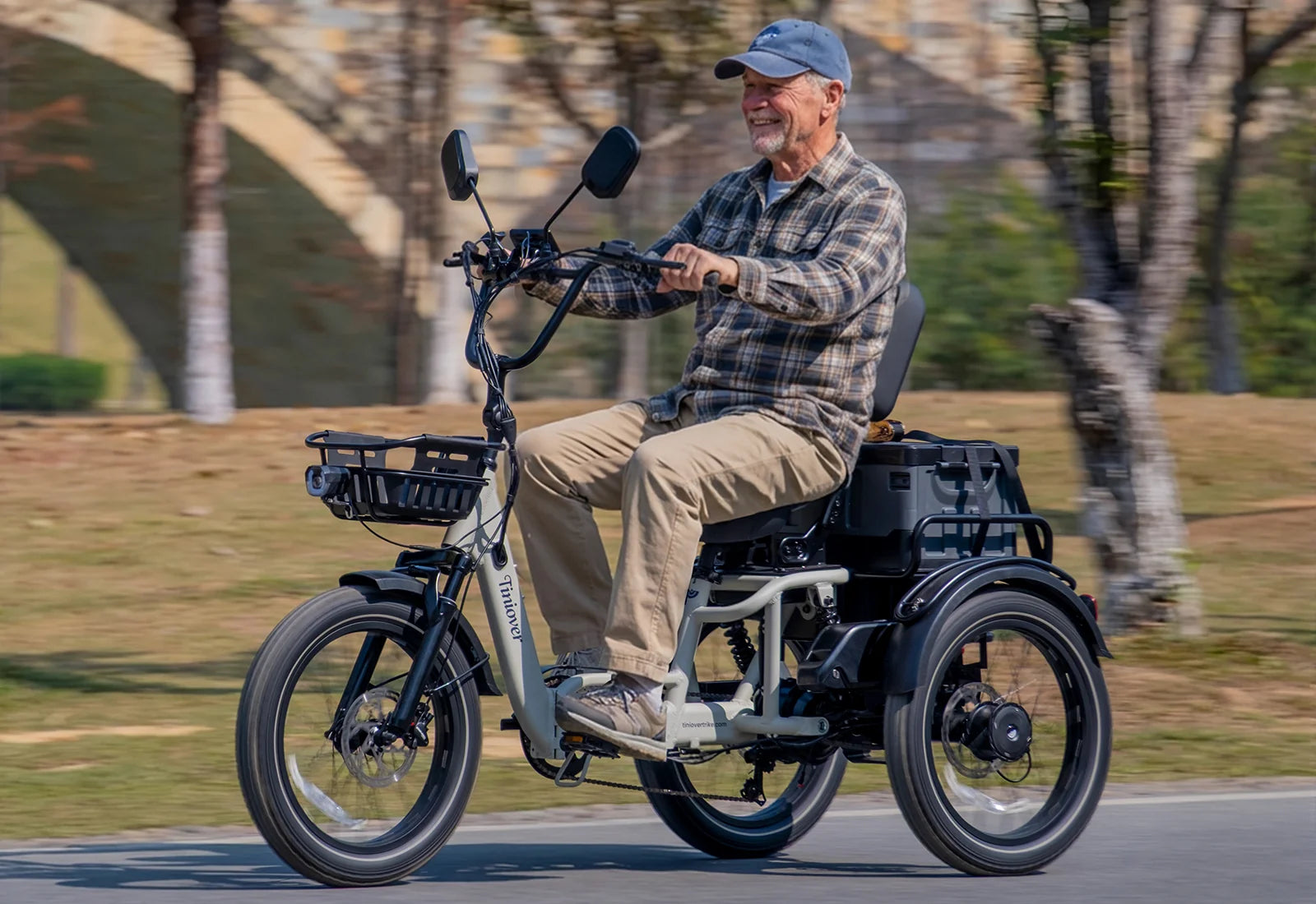 White Tiniover River electric trike with a rider, front basket, rear storage box, and fat tires on a paved path